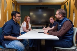 Four colleagues engaged in a discussion around a white table, with smartphones placed on the table, in a modern conference room.